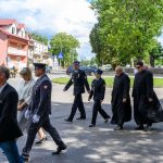 Grupa ludzi w eleganckich ubraniach idzie ulicą. W tle drzewa, czerwony budynek i kościół z cegły. Niebo zachmurzone.