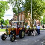 Traktor z ozdobną przyczepą na ulicy, z tyłu ceglana budowla. Ludzie w strojach regionalnych, wokół zielone drzewa.