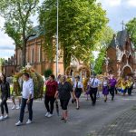 Procesja ludzi idących przed ceglaną świątynią na tle zieleni. Uczestnicy niosą wieńce dożynkowe. Pogodne niebo w tle.