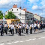 Orkiestra maszeruje ulicą w słoneczny dzień, w tle niskie budynki i drzewa. Uczestnicy w białych koszulach i czapkach. Niebo z chmurami.