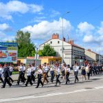 Parada orkiestry dętej na ulicy, w tle budynki i niebo z białymi chmurami. Uczestnicy w uniformach maszerują obok billboardu. Ciepły, słoneczny dzień.