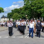 Grupa osób maszeruje w wielobarwnych mundurach z instrumentami dętymi, na tle zielonych drzew i niebieskiego nieba.