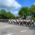 Orkiestra maszeruje ulicą w słoneczny dzień. Muzycy w białych koszulach i czapkach grają na instrumentach dętych. W tle zielone drzewa i niebo z chmurami.