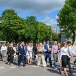Grupa ludzi na ulicy w słoneczny dzień, trzymają flagi i chleb. Tło stanowią drzewa i niebieskie niebo z chmurami. Uczestnicy są elegancko ubrani.