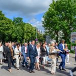 Grupa ludzi ubrana elegancko idzie ulicą, trzymając chleb na białych serwetkach. Tło to zielone drzewa i niebo z chmurami. Po lewej stronie widać traktor z ozdobami.