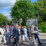 Grupa ludzi w eleganckiej odzieży idzie ulicą na tle zielonych drzew. Jasne niebo z chmurami, po prawej strony tablice ogłoszeniowe. Osoba niesie tradycyjny chleb na białym obrusie.