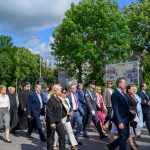 Grupa ludzi w eleganckich strojach i mundurach idzie ulicą. Tło to zieleń drzew, niebo częściowo pochmurne. Po prawej widoczne są plakaty.
