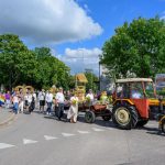 Grupa ludzi w tradycyjnych strojach idzie za czerwonym traktorem ciągnącym przyczepę z kwiatami. Tło z zielonymi drzewami i niebieskim niebem. Spokojna scena festiwalowa na ulicy.