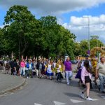 Grupa ludzi uczestniczy w procesji ulicznej, niosąc ozdobną ramę z obrazem Maryi. Tło to zielone drzewa i błękitne niebo z chmurami. Dominuje kolor zielony, szary i różne kolory ubrań.