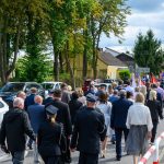 Grupa ludzi w eleganckich ubraniach idzie ulicą w uroczystym pochodzie. Na tle zielonych drzew widoczne są kolorowe flagi. Pogodne niebo z białymi chmurami.