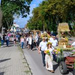 Parada dożynkowa na drodze, ludzie w odświętnych strojach, kolorowe dekoracje. Drzewa w tle, jasno i słonecznie. Traktor z wieńcem z przodu.