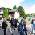 Grupa ludzi idzie ulicą w eleganckich strojach. Tło to domy z zielonymi ogrodami i jasne niebo. Na czele osoba w mundurze.