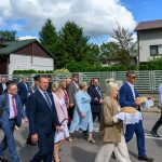 Grupa elegancko ubranych ludzi idzie ulicą, niosąc chleb na białym obrusie. Tło to domy i zielone drzewa. Pogoda słoneczna, niebo z chmurami.