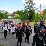 Na zdjęciu widoczny jest pochód strażaków w czarnych mundurach z chorągwiami. W tle znajdują się zielone drzewa i domy z czerwonymi dachami. Na niebie widać kilka białych chmur.
