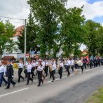 Grupa muzyków maszeruje wzdłuż ulicy z drzewami w tle. Ubrani w białe koszule i czarne spodnie grają na instrumentach dętych. W tle widać zielone drzewa i budynek z czerwonym dachem.