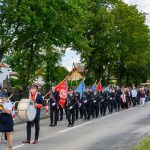 Parada na ulicy z ludźmi w mundurach, orkiestra z instrumentami i kolorowe sztandary. Drzewa w tle, zielone liście. Pogoda pochmurna.