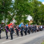 Ulicą maszeruje grupa osób w mundurach z kolorowymi flagami. W tle drzewa oraz plac zabaw. Kolory żywe, dominują zieleń i czerń.