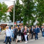 Grupa ludzi spaceruje ulicą przy zielonych drzewach i budynku z czerwonym dachem. Mają eleganckie stroje. Tło ożywia plac zabaw.