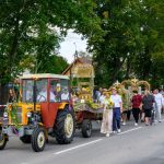 Parada z traktorem w wiejskim stylu, kolorowy wóz z kwiatami, obok tłum ludzi, zielone drzewa w tle.