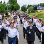 Orkiestra dęta maszeruje ulicą w białych koszulach i czapkach, grając instrumenty. W tle zielone drzewa i znaki drogowe. Okolica jest miejska z niską zabudową.