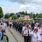 Pochód orkiestry dętej i strażaków w mundurach idących ulicą. Domy i zieleń w tle, jasne niebo z chmurami. Uczestnicy niosą instrumenty i flagi.