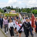 Grupa ludzi w marszu po ulicy, niektórzy z flagami. Ubrani formalnie, w tle zieleń i zabudowania. Pogoda pochmurna.
