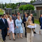 Grupa ludzi w eleganckich strojach idzie ulicą. Kobieta i mężczyzna niosą chleby na białych serwetkach. W tle zielone ogrody i domy.
