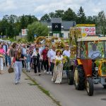 Grupa ludzi idzie ulicą obok ozdobnego traktora z wieńcem. Tło to zielone drzewa i szare niebo. Dominuje kolorowa procesja.