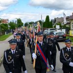 Parada strażaków w czarnych mundurach z odznaczeniami i sztandarami na wiejskiej ulicy. Tło stanowią domy i drzewa. Niebieskie niebo z chmurami.