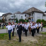 Orkiestra strażacka w białych i czarnych strojach maszeruje ulicą. W tle są domy i zachmurzone niebo. Towarzyszy im grupa mieszkańców.