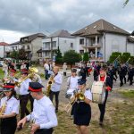 Orkiestra maszeruje ulicą przed białymi budynkami. Ubrania muzyków w bieli, czerni i czerwieni. Tło to chmury i widzowie.