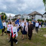 Grupa młodzieży w białych koszulach niesie sztandary na tle domów i chmur. Tło jest zielone i pochmurne. Eleganckie ubrania wskazują na uroczystość.