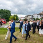 Grupa ludzi idzie po trawiastej ścieżce; na czele mężczyzna niesie chleb. W tle stoją domy i czerwony wóz strażacki. Pochmurne niebo.