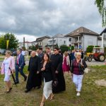Grupa ludzi w formalnych strojach idzie wzdłuż trawiastej drogi. W tle widać budynki mieszkalne i traktor. Niebo jest zachmurzone.