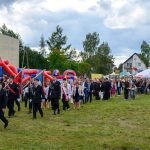 Na zdjęciu widać paradę z pocztem sztandarowym na tle zieleni i barwnych namiotów. Niebo jest pochmurne, a w tle widoczne są budynki. Uczestnicy ubrani są odświętnie, a teren jest trawiasty.