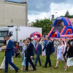 Grupa ludzi w eleganckich strojach idzie po trawie na tle kolorowego dmuchanego toru przeszkód. Nad nimi chmury. W tle widoczny biały samochód dostawczy.