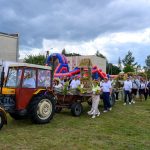 Na zdjęciu widoczny jest kolorowy festyn z czerwonym ciągnikiem i platformą z ozdobami dożynkowymi. W tle znajdują się dmuchane atrakcje i zabudowania. Nad sceną unosi się pochmurne niebo.