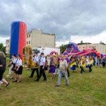 Grupa ludzi na polu niesie dekoracje z kłosami zboża. Tło stanowi budynek i kolorowe dmuchańce. Na niebie wiszą ciemne chmury.