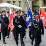 Grupa strażaków w czarnych mundurach maszeruje z kolorowymi sztandarami. W tle budynek i ludzie. Dominuje czerń i czerwień.