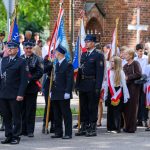 Grupa strażaków i młodzieży z flagami stoi na ulicy. Mundury są ciemne, a tło stanowi ceglana budowla i zieleń. Uroczystość odbywa się w pogodny dzień.