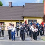 Parada orkiestry dętej w mundurach maszeruje ulicą. Jasne, letnie słońce, kolorowe flagi w tle. Domy w tle mają dachy w odcieniach brązu.
