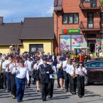 Orkiestra dęta maszeruje ulicą w słoneczny dzień. Członkowie noszą mundury w odcieniach bieli, granatu i czerwieni. W tle żółty budynek i czerwony sklep groszek.