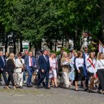 Grupa ludzi na paradzie, ubrani elegancko, niosą flagi i chleb. Tło zielone drzewa, słoneczny dzień. Uroczysta atmosfera.