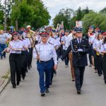 Orkiestra strażacka maszeruje ulicą, ubrane w biało-niebieskie mundury. Tło to zielone drzewa i kilka widzów. Instrumenty to trąbki i klarnety.