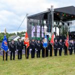 Grupa strażaków w mundurach i czapkach stoi na trawie z flagami. Na tle sceny z czarnym dachem i sprzętem, niebo pochmurne. Zgromadzenie formalne.