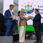 Grupa osób w eleganckich strojach na scenie wymienia się chlebem. Tło to jasny ekran z kolorowym logo. Atmosfera oficjalna.