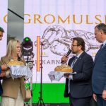 Cztery osoby na scenie prezentują tradycyjne chleby na białych serwetach. W tle widoczny jest baner z napisem „Gromulska”. Sceneria jest kolorowa i uroczysta.