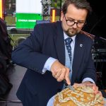 Mężczyzna w garniturze kroi ozdobny chleb. Tło stanowią kolorowe światła i zielona tablica. Ujęcie sceniczne na świeżym powietrzu.