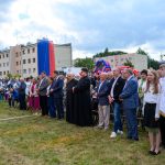 Grupa ludzi uczestniczy w plenerowym wydarzeniu. W tle widoczne są budynki i kolorowe dmuchańce. Niebo jest pochmurne.
