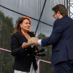 Kobieta w czarnym żakiecie trzyma mikrofony, mężczyzna podaje jej chleb na białym talerzu. Tło sceniczne z drzewami.